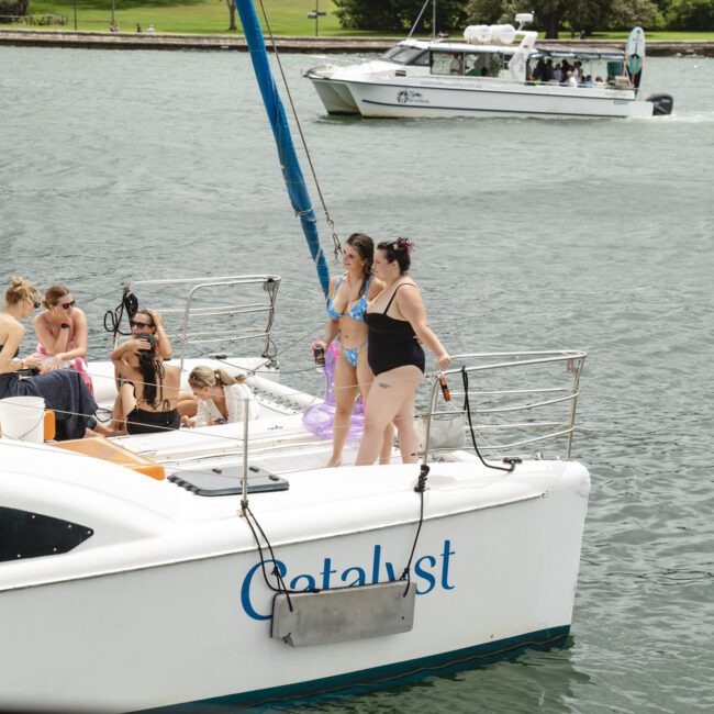 People are enjoying a day on a white sailboat named "Catalyst" on a lake. Some are lounging under the sun, while others stand near the edge. Another boat is visible in the background on the water.