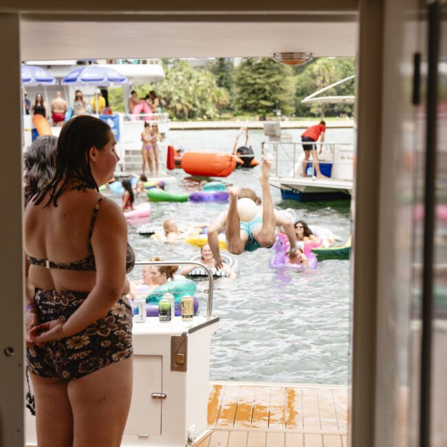 A woman in a patterned bikini stands on a boat deck, watching people enjoying a day at the lake. Some are swimming and others are using inflatables. A man is diving into the water. Trees and another boat are visible in the background.