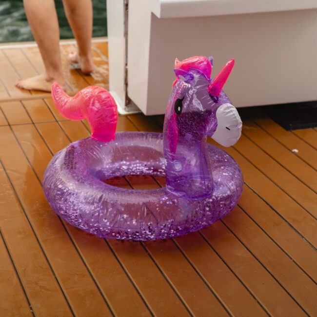 A purple inflatable unicorn float with a shiny mane and tail is placed on a wooden deck near the water. A person with bare feet is partially visible in the background.