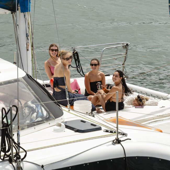 Four people are sitting on the deck of a sailboat, smiling and enjoying the day. The boat is on calm water, with blue sail covers visible. The scene conveys a relaxed, sunny atmosphere.