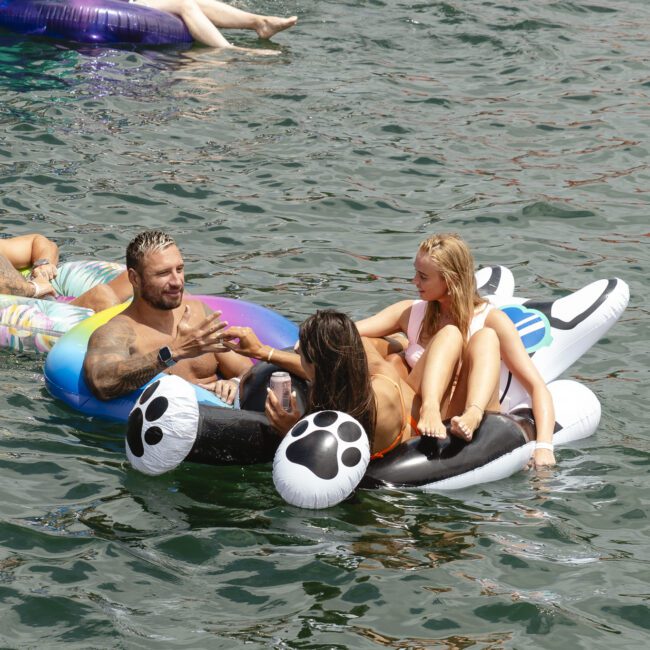 A group of four people relax on inflatable floats in the water. Two men and two women are chatting and enjoying the sunny day on inflatable rafts shaped like a paw print and a colorful ring. The surface of the water is visible and rippling.