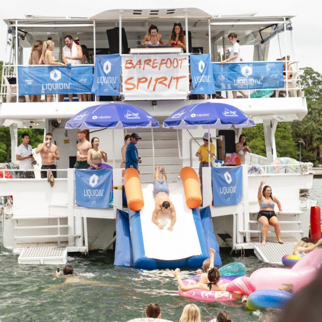 A multi-deck party boat on the water with people enjoying a sunny day. A person slides down a water slide into the water while others swim and relax on colorful floaties. Signs read "Barefoot Spirit" and "LIQUID." Trees and skyscrapers are in the background.