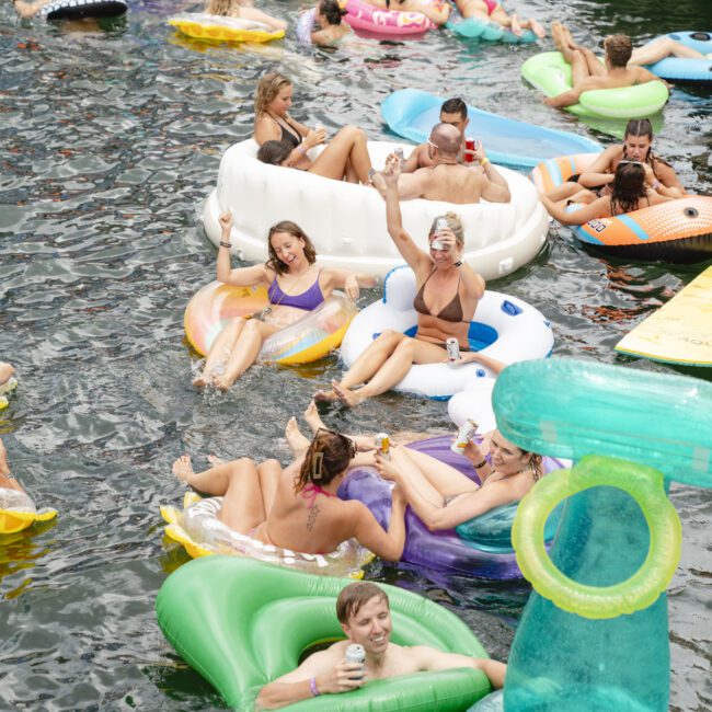 A group of people enjoying a day on the water, lounging on various colorful inflatable floats. They appear relaxed and cheerful, holding drinks, with more floats and a boat visible in the background.