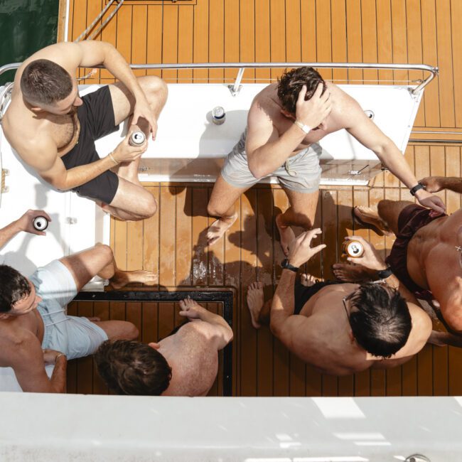 A group of shirtless men are gathered on a boat deck, holding cans of drinks. They appear to be having a casual conversation, surrounded by wooden flooring and the green water of a marina or dock area.