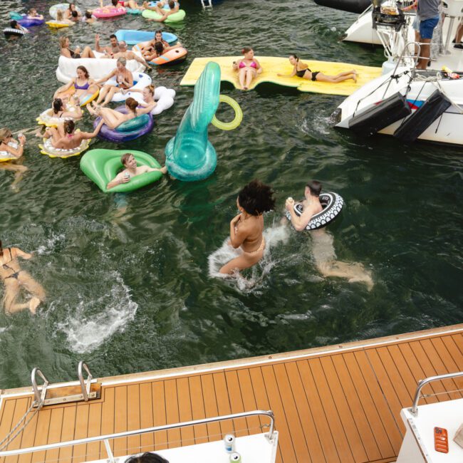 People enjoy a lively day on the water, swimming and lounging on inflatable floats of various shapes. A boat is docked nearby with more people mingling. The scene is festive and relaxed.