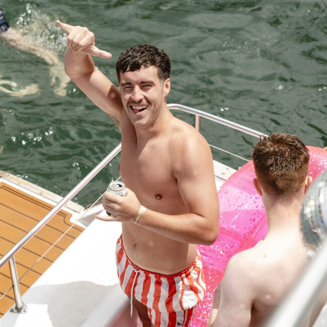 A person in red and white striped swim trunks stands on a boat, making a hand gesture. They are smiling and holding a beverage can. Another person is partially visible beside them, and there's a pink float in the water.