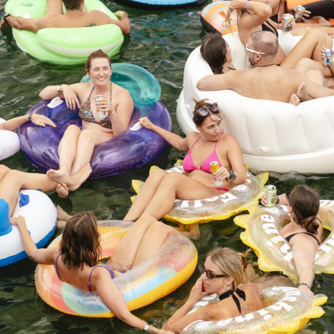 A group of people relax on colorful inflatable pool floats in water, holding drinks and chatting. The floats are shaped like rings and animals, including a swan. The scene is lively and festive, with people enjoying a sunny day.