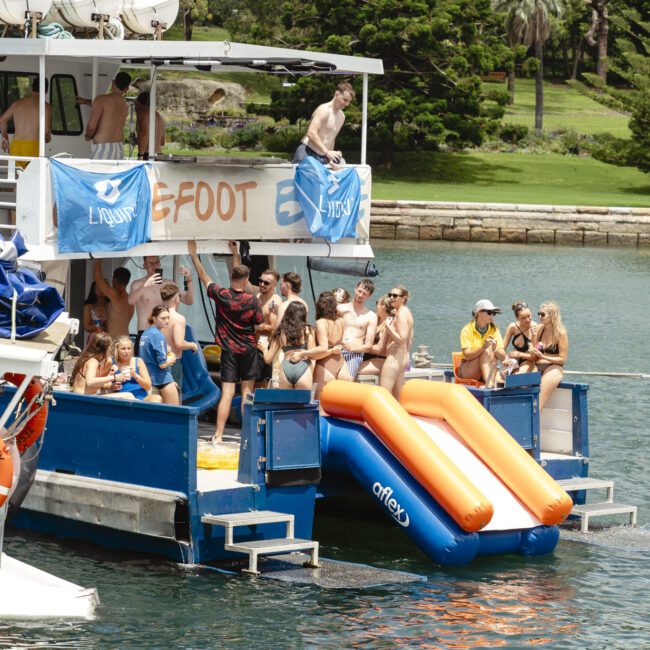 A group of people enjoying a sunny day on a boat equipped with inflatable slides leading into the water. Some are sitting while others are standing and talking. The boat is docked near a grassy shoreline with lush greenery in the background.