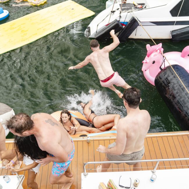 A group of people enjoying a sunny day on a boat. One person is jumping into the water near a large pink inflatable unicorn. Others lounge on floaties and sit on the boat's deck, surrounded by water.