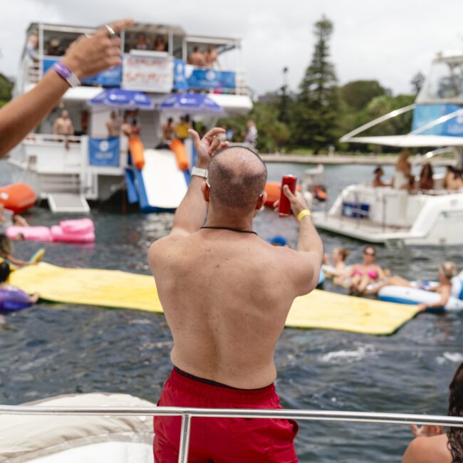 A man in red shorts holds a drink and gestures towards a crowd of people enjoying a boat party. The scene includes multiple boats, people on inflatables in the water, and a festive atmosphere under a partly cloudy sky.