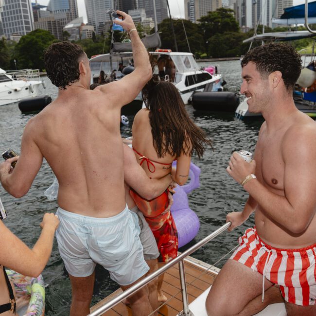 People enjoy a boat party, some in swimsuits holding drinks. Boats and a city skyline are visible in the background. A person in striped shorts stands on deck while others are in the water with inflatable toys.