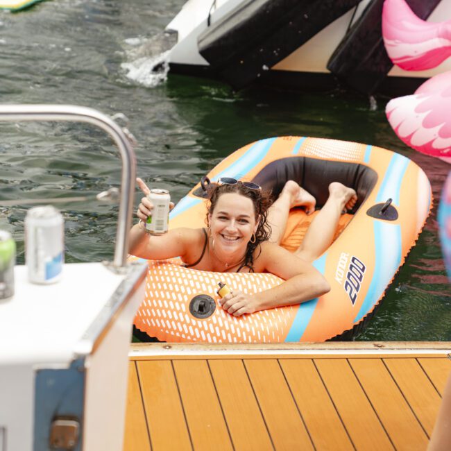 A woman smiles and holds up a canned drink while lying on an orange inflatable float in the water. She is next to a dock, and a large inflatable pink flamingo is partially visible in the background.