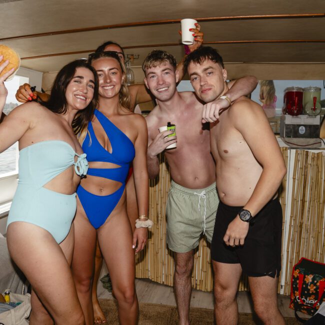 A group of five young adults posing on a boat, smiling. One woman is in a blue swimsuit, holding a drink, while another has a light blue bikini and holds up a bun. Three men stand with them, casually dressed. The interior is decorated with bamboo accents.