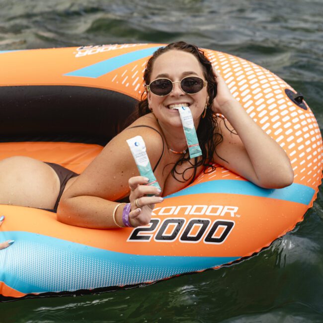 A person lounges on an orange inflatable raft labeled "CONDOR 2000" in the water. They are wearing sunglasses, smiling, and holding two rolled-up items. Another hand rests on the raft.