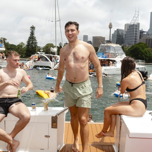 A group of people enjoying a sunny day on a boat. One man stands smiling, while others sit and chat. Boats and a city skyline with tall buildings are visible in the background, set against a partly cloudy sky.