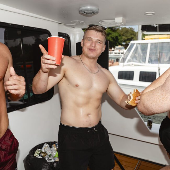 A group of shirtless people on a boat are enjoying food and drinks. One person in the center holds a red cup and poses while another holds a sandwich. There's a bin with trash in the background alongside windows and a view of the water.