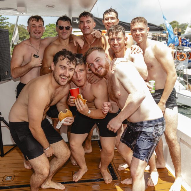 A group of nine men in swimsuits pose together on a boat deck. Some are holding drinks and one has a sandwich. In the background, there's water with other boats and inflatable structures visible. Everyone appears to be enjoying a sunny day outdoors.