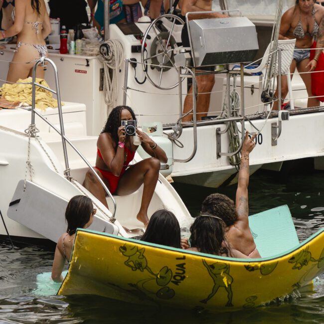 People are enjoying a sunny day on a boat. A person in a red swimsuit is taking a photo from the slide. Others are relaxing on floats in the water, socializing and having fun. A man is cooking on a grill.