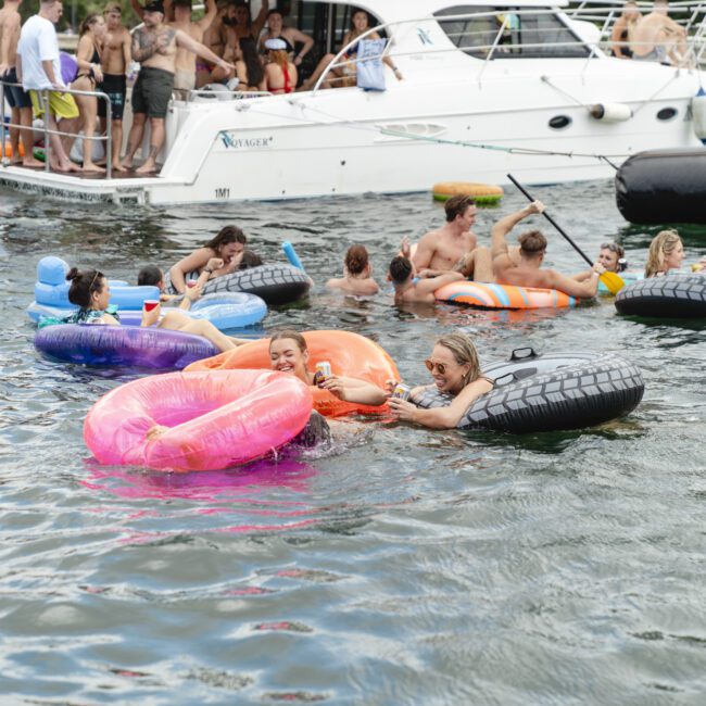 People enjoying a sunny day on the water, floating on colorful inflatable rings near a docked boat. Some are swimming, while others relax on the boat or sit on inflatables. The scene is lively and festive.