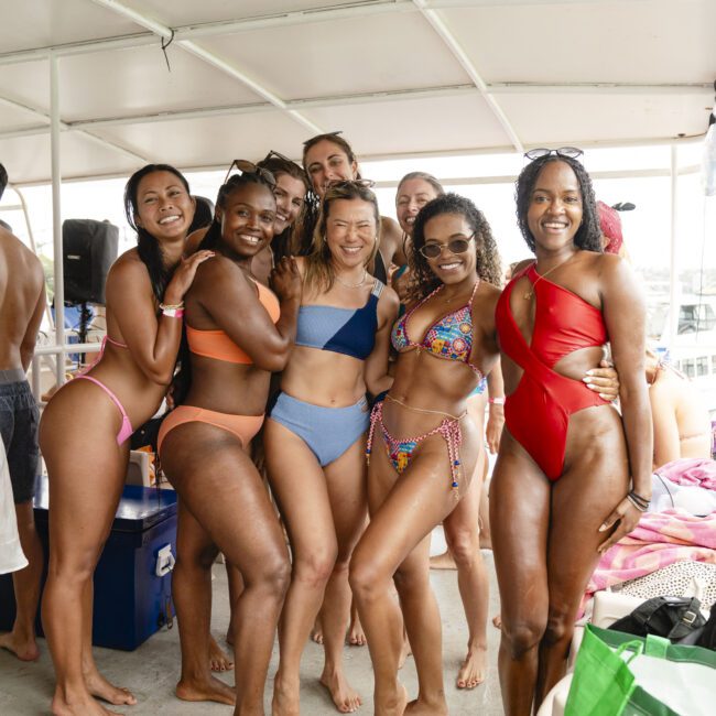 A group of people in swimsuits smile and pose together on a boat. The sun is shining, and everyone looks relaxed and happy. The interior of the boat has a white ceiling and various items scattered around.