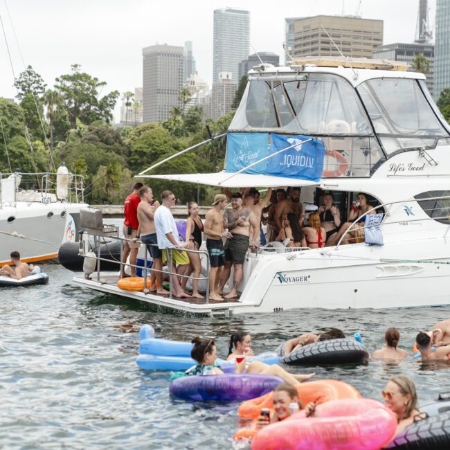 A group of people enjoying a party on a docked white yacht surrounded by inflatables in the water. Some are in swimsuits, others are lounging on floaties. City buildings and trees are visible in the background under a partly cloudy sky.