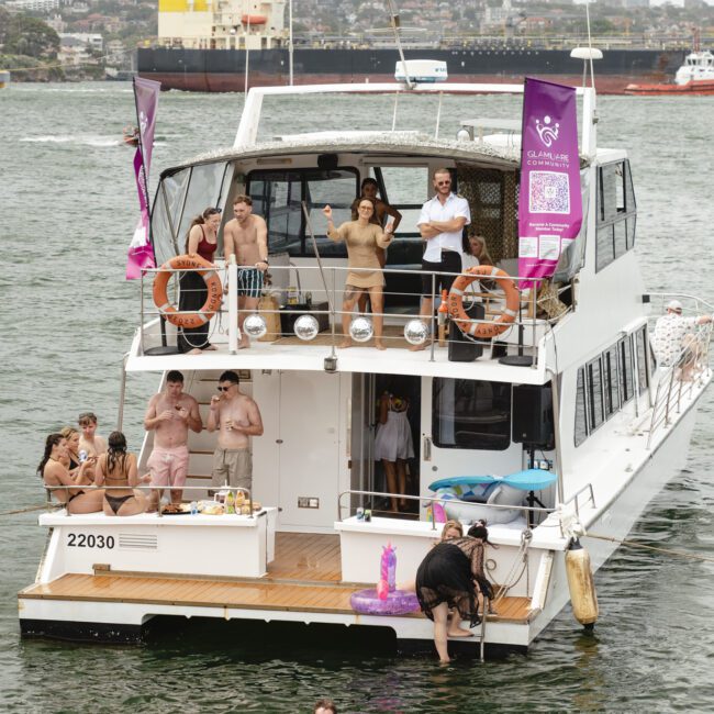 A group of people enjoying a party on a white boat on the water. Some are standing on the upper deck, and others are gathered on the lower deck with a food table and pink pool toys. A few large ships are visible in the background.