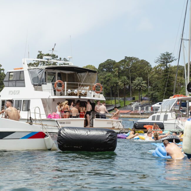 A group of people are gathered on and around several boats in a harbor. Some are on inflatable floats in the water. Trees and a grassy area are visible in the background under a partly cloudy sky.