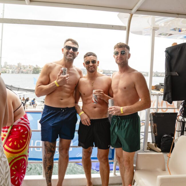 Three men in swim trunks stand on a boat, each holding a drink. They are wearing sunglasses and wristbands. The background shows water and distant land. A person in swimwear is partially visible on the left.