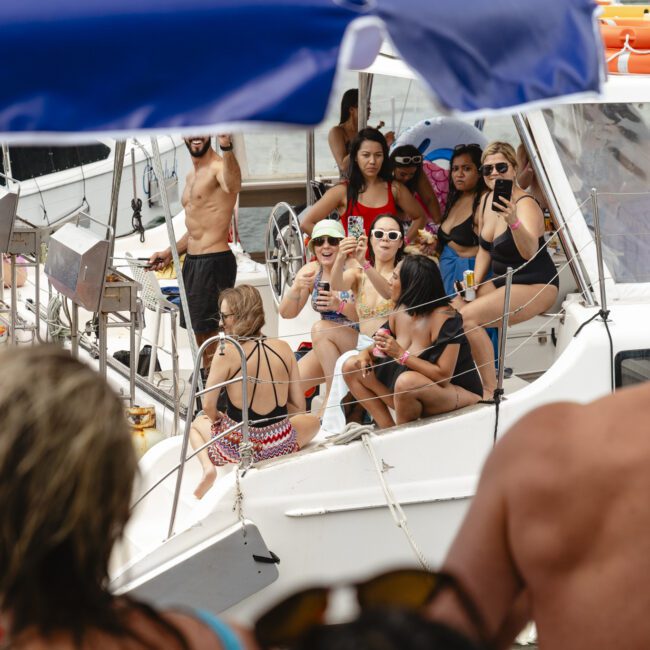 A group of people in swimwear is enjoying a sunny day on boats. Some are sitting and chatting, while others stand and look around. A large inflatable flamingo floats nearby. The atmosphere is lively and festive.