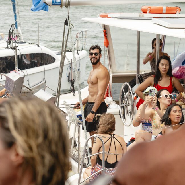 People enjoying a sunny day on a boat, some taking photos and relaxing. A man in swimwear stands at the center, smiling. The background shows additional boats and water, suggesting a social gathering or party on the water.