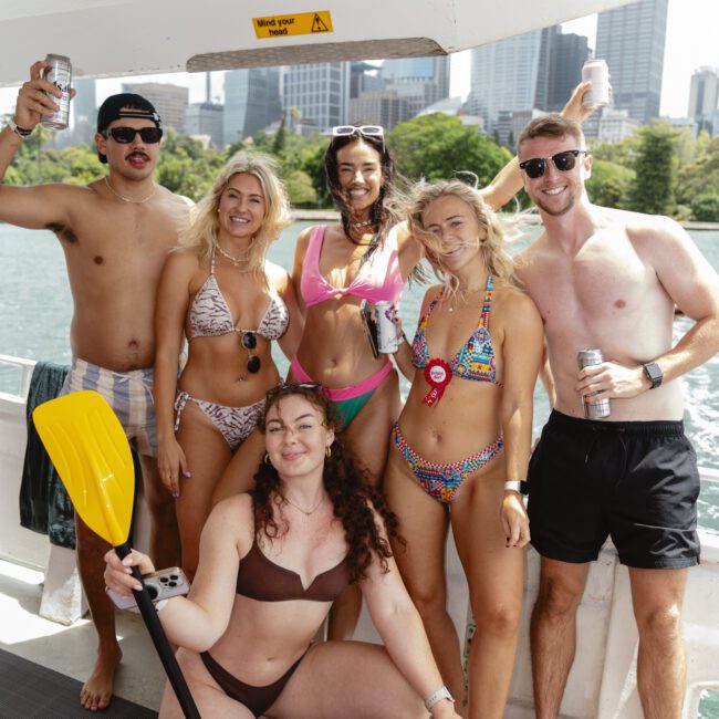 A group of six young adults in swimwear smiling and posing on a boat. They are holding drinks and a paddle, with a city skyline and greenery visible in the background. The atmosphere is cheerful and festive.