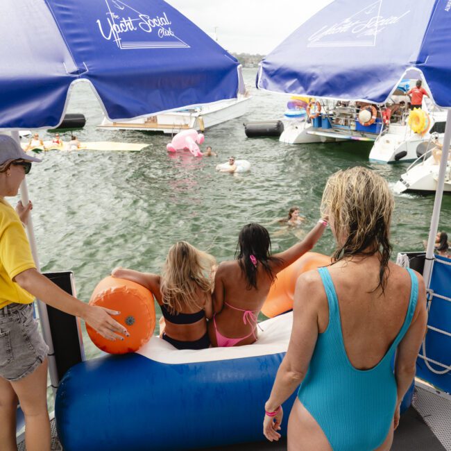 People enjoying a floating water park with inflatable structures on a body of water. A lifeguard in yellow supervises. Two women and a child prepare to slide into the water. Blue umbrellas provide shade nearby.