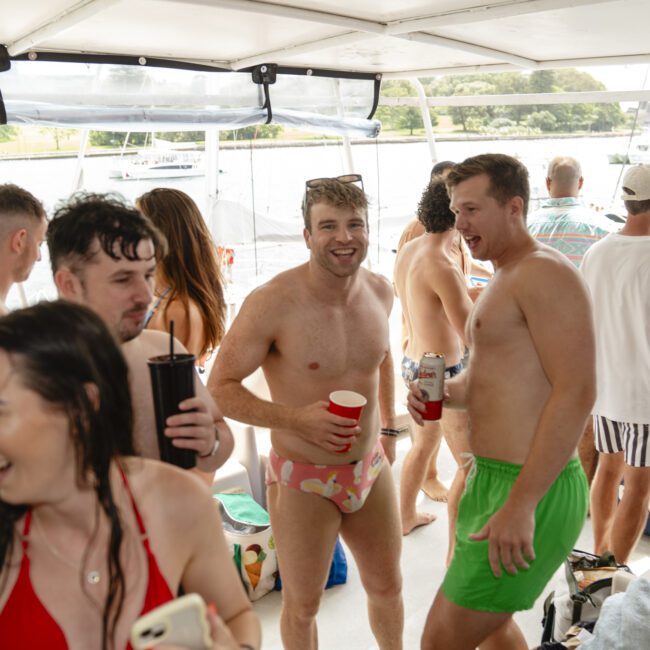 A group of people in swimwear enjoying a boat party. Some hold drinks while others converse. The background shows water and other boats. Smiles indicate a fun and relaxed atmosphere on a sunny day.