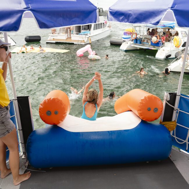 Person in a swimsuit preparing to jump off a floating platform into the water, surrounded by boats and people swimming. A person in a yellow shirt and hat stands nearby. Blue and white canopy above.