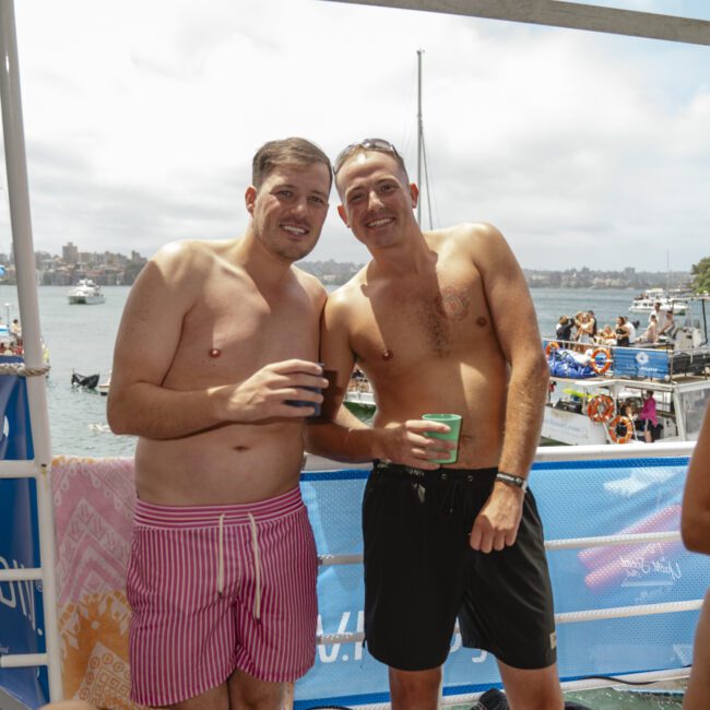 Two men in swimwear stand on a boat holding drinks, smiling at the camera. The background shows a harbor with boats on the water and a cloudy sky.