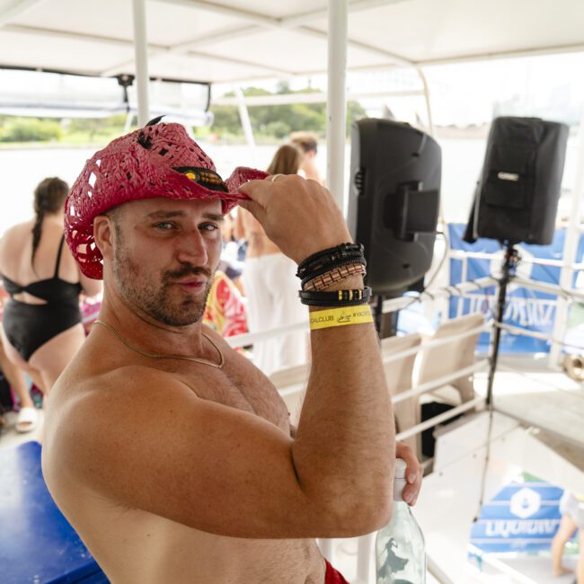 A man wearing a pink cowboy hat poses confidently on a boat deck filled with people. He's shirtless, wearing multiple bracelets, and holding onto the hat's brim. Other passengers in swimwear are seated and standing in the background.