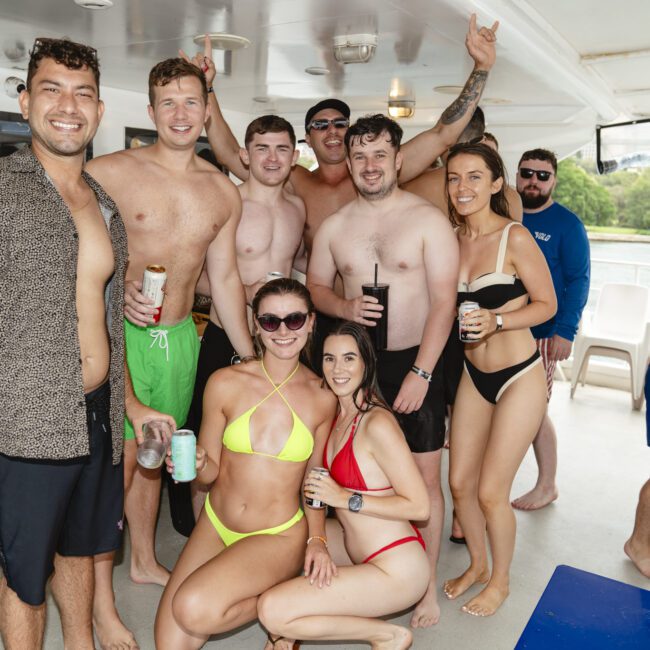 A group of people in swimsuits are posing and smiling on a boat. Some hold drinks, and one person gives a thumbs-up. Trees and water are visible in the background.