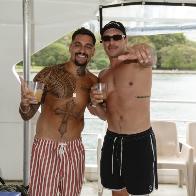 Two men on a boat smiling and holding drinks. The man on the left has tribal tattoos and wears red and white striped shorts. The man on the right wears sunglasses, a cap, and black shorts, pointing toward the camera. Trees and water are in the background.