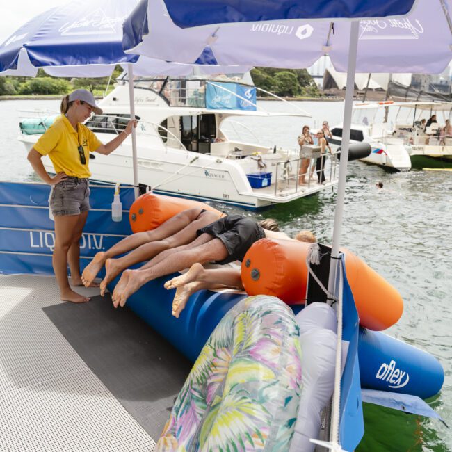 People lying on inflatable tubes on a dock near the water, with a person standing nearby. Blue and white umbrellas provide shade. A floral-patterned float is in the foreground, and boats are visible in the background.