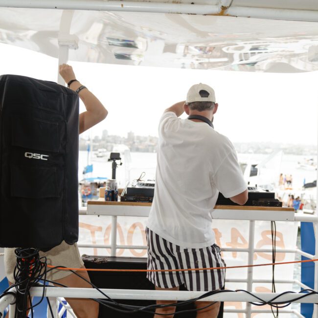 Two people on a boat are DJing at an outdoor event. One is wearing a white shirt and striped shorts. A speaker is visible next to them. The sea and several other boats are in the background.