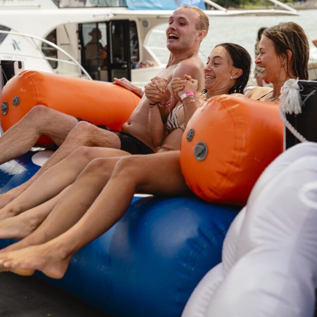 Four people are sitting on an inflatable water toy attached to a boat. They are laughing and holding hands, wearing swimsuits. Other boats are visible in the background, and the water is calm.