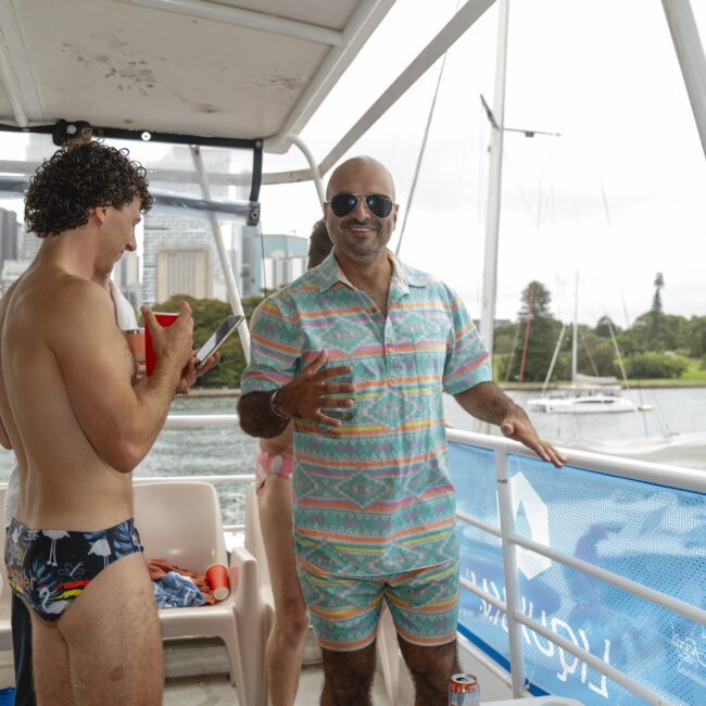 Two people on a boat wearing swimwear. One is in trunks holding a cup, the other in a colorful patterned outfit with sunglasses, smiling. A body of water and cityscape are visible in the background.