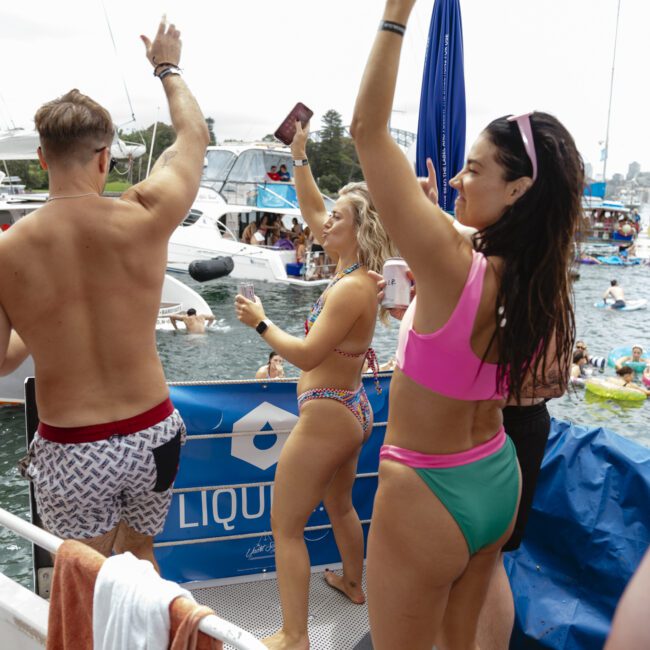 People in swimsuits are dancing and celebrating on a boat during a sunny day. Other boats and people in the water are visible in the background. There are festive decorations, and the atmosphere is lively and joyful.