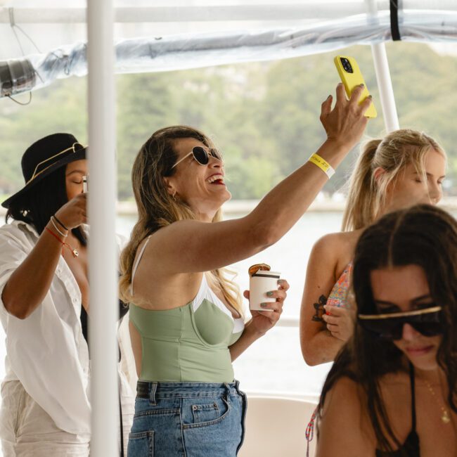 A group of women enjoying a boat outing. One woman in a green swimsuit and sunglasses is raising her phone for a photo, holding a cup. Others are dressed casually, socializing, with overcast skies and water in the background.