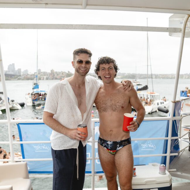 Two men on a boat deck, smiling. The man on the left wears sunglasses and a light shirt, holding a drink can. The man on the right is shirtless, wearing swim trunks and holding a red cup. Boats and water are visible in the background.