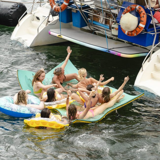 A group of people relax on floating mats in the water near a docked boat. Some are lounging, while others are sitting up, engaging with each other. They're surrounded by inflatable rings and the rear of a boat with steps leading down.