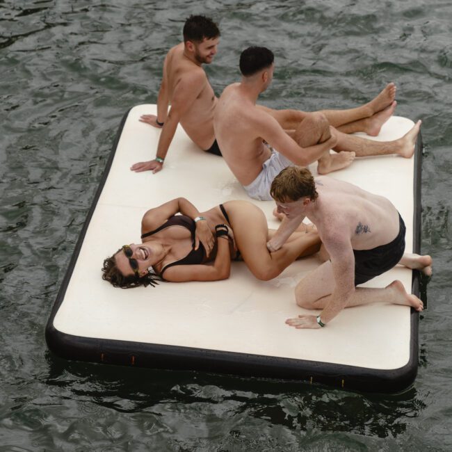 Four people are lounging on a large floating mat in the water. Two men are sitting upright, one man is reclining at the back, and a woman is lying down with sunglasses on, smiling and relaxed. Waves ripple around them.