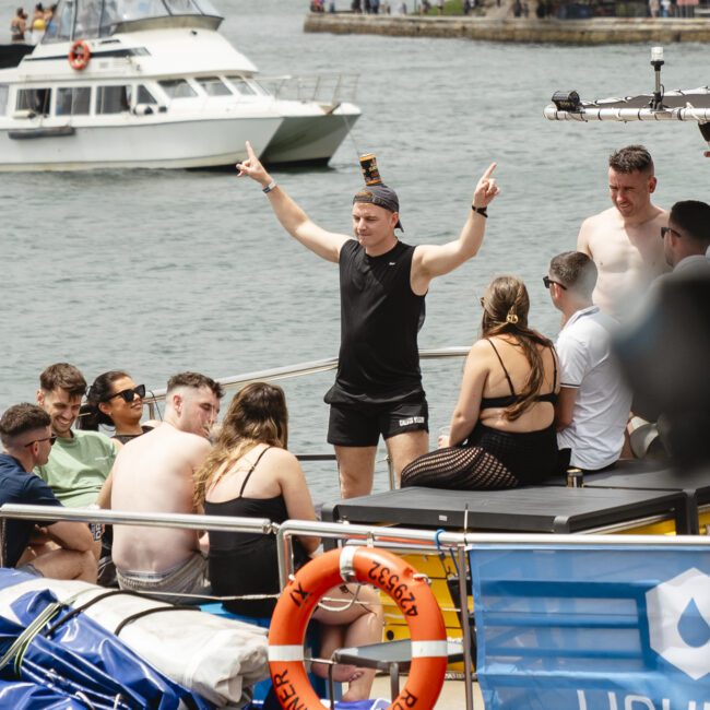 A person in a black shirt and shorts stands on a boat, making peace signs. They are surrounded by others sitting and talking. The background features a waterway, a docked boat, and distant hills. A life preserver and equipment are visible on the boat.