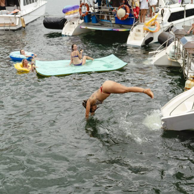 A person in a swimsuit is diving off a boat into the water, surrounded by others on floats and swimming nearby. Several boats are anchored, with people observing the scene and enjoying the water.