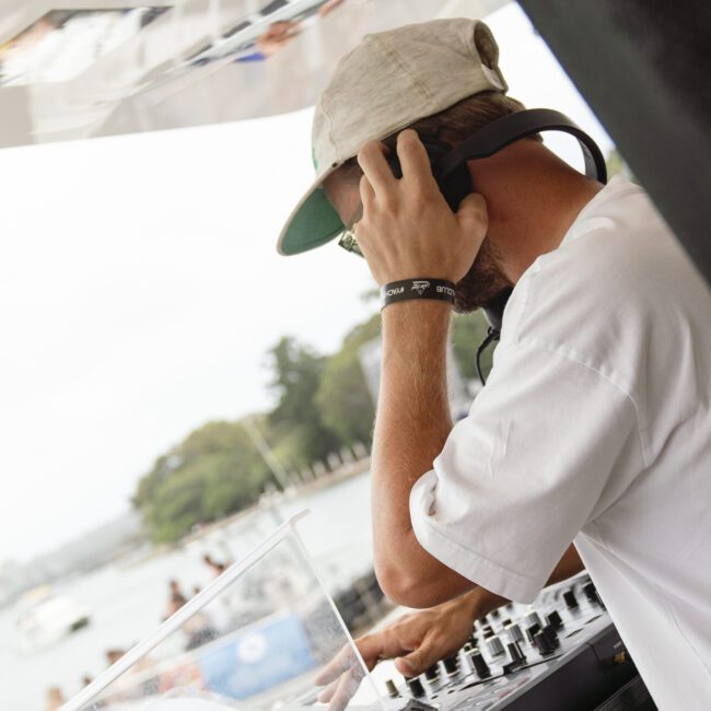 A DJ wearing a white shirt and cap uses turntables at an outdoor event. He's adjusting headphones with one hand. The background shows trees and a body of water, with people enjoying the scene.